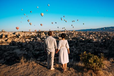 Hot air balloon flying over spectacular Cappadocia - couple watching hot air balloons at the hill of Cappadocia. Couple standing by each other in Turkey Cappadocia with hot air balloons at sunrise.